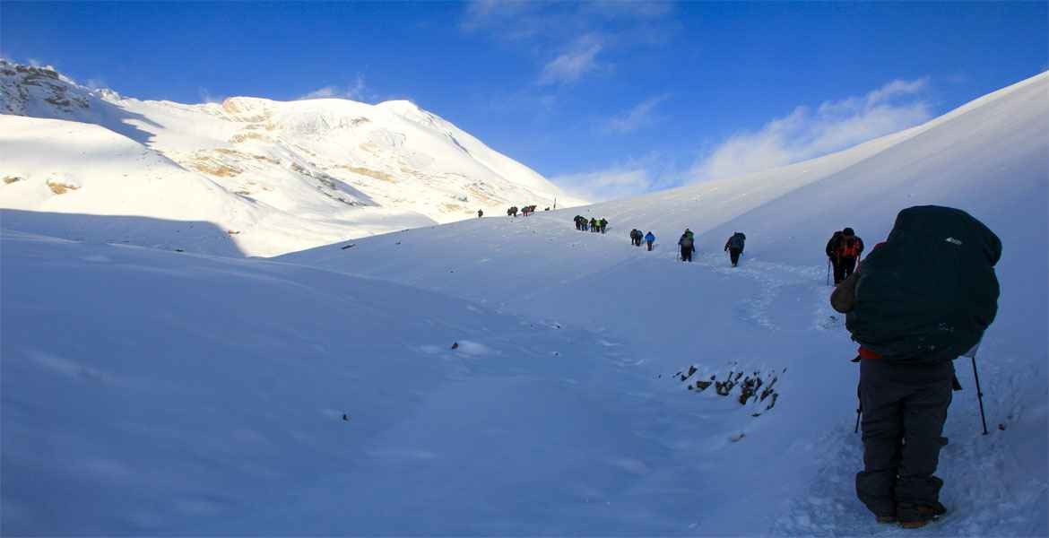 trekkers are crossing thorong la early morning wearing warm trekking gear, snow on the trail, the blue sky, and thorong peak.